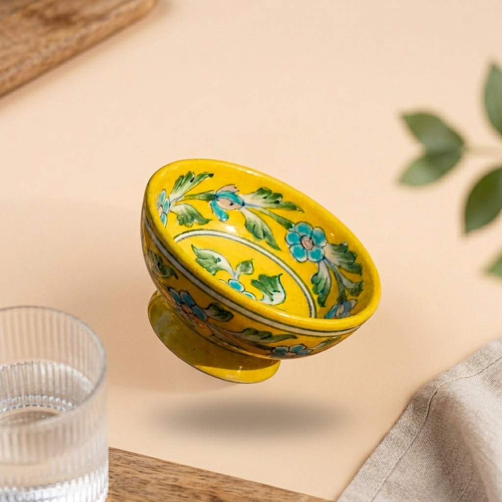 Handcrafted Jaipur Blue Pottery footed bowl in yellow displaying hand-painted blue floral and green vine motifs on a beige surface