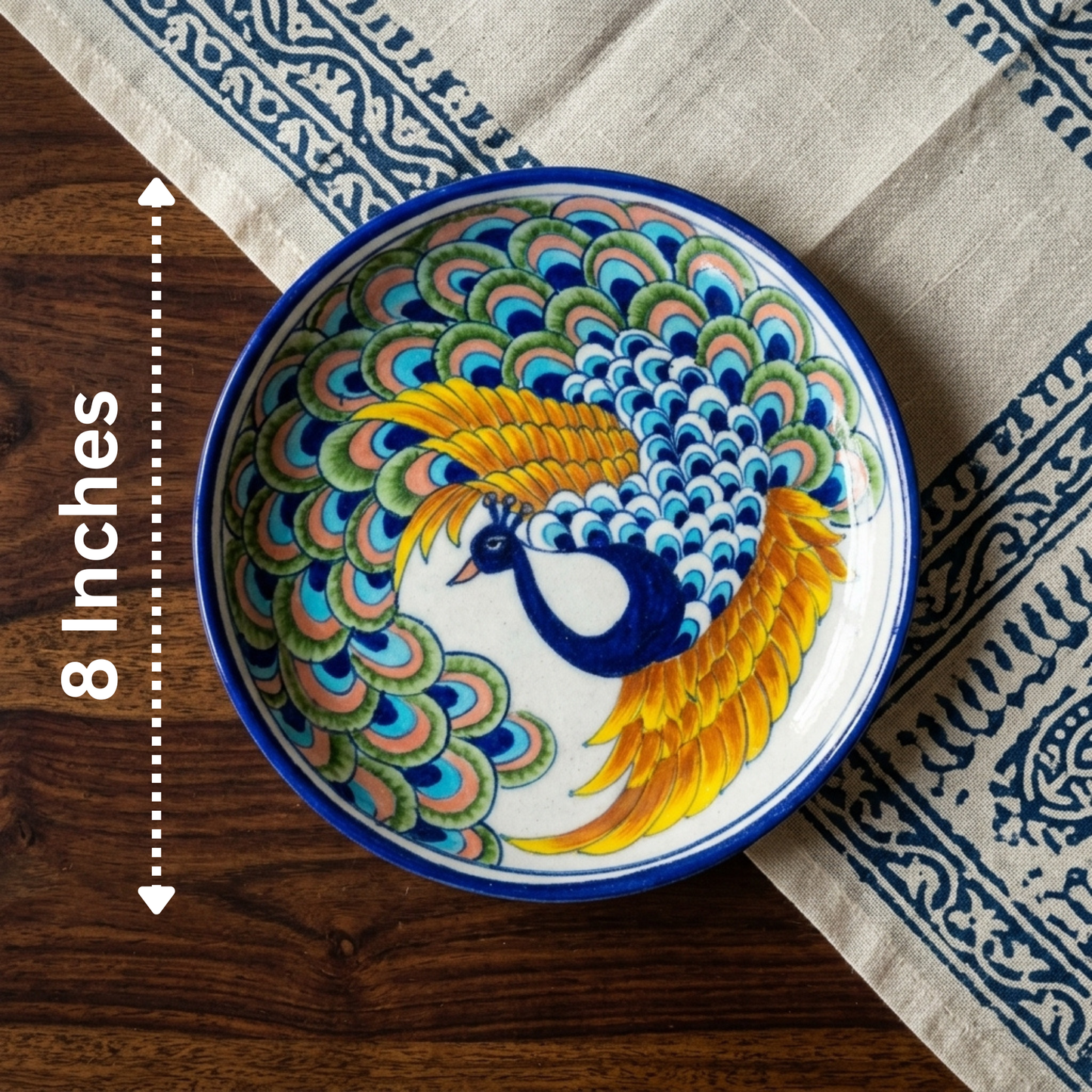 Hand-painted 8-inch Jaipur Blue Pottery plate with cobalt peacock and golden feather motif on block-print fabric.