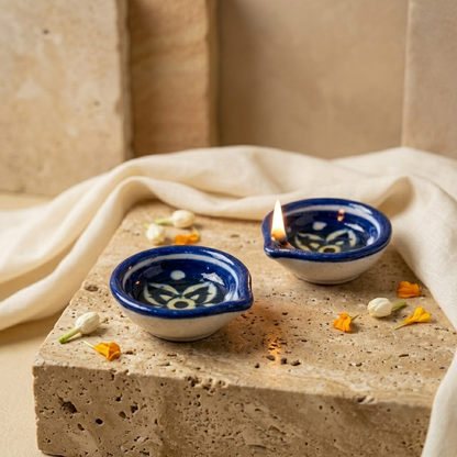 Set of two handcrafted Jaipur Blue Pottery diyas styled on natural stone with soft linen fabric and scattered flower petals
