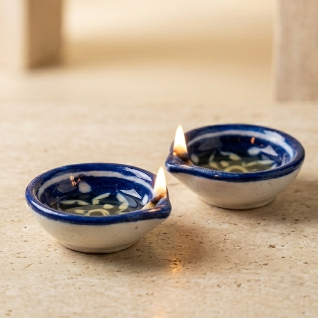 Two hand-painted Blue Pottery diyas on a stone surface with one diya beautifully lit, warm festive decor